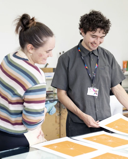 Two people examining orange geometric prints on a table in an art studio, with another person working in the background. Two people examining orange geometric prints on a table in an art studio, with another person working in the background.